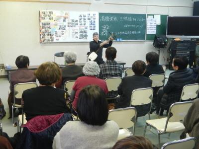 津軽三味線を手に話をする高橋さんとその話を聞く参加者たちの写真