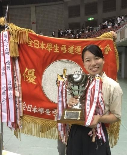 第66回全日本学生弓道選手権大会において団体戦で優勝し、会場で優勝旗を背にして優勝カップを持つ小川結花さんの写真